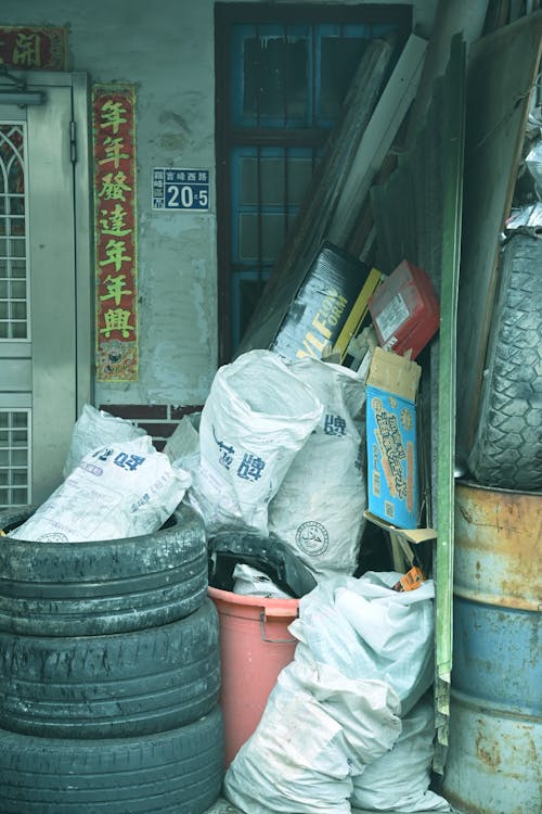 Grand tas de déchets et pneus à l’extérieur d’un bâtiment, sacs blancs et conteneurs colorés empilés près d’une porte. code couleur des conteneurs poubelles